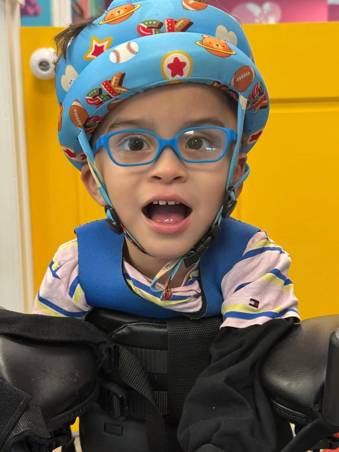 A child in a supported walker, wearing blue glasses and a colorful helmet